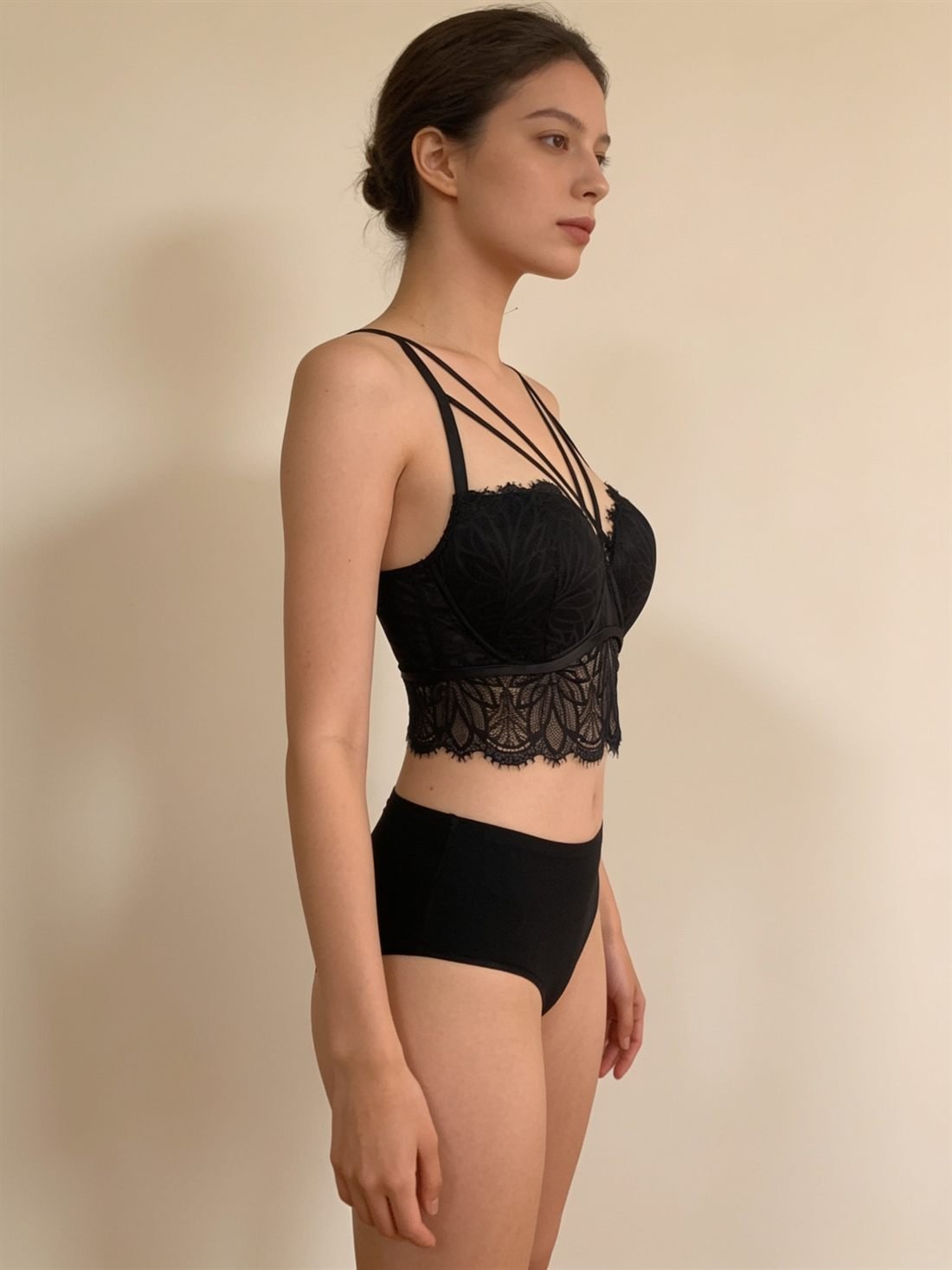 Manken üzerinde tam yandan profil görünüm; kesim ve boy — Siyah dantel çapraz askı büstiyer; göğüs üzerinde çapraz saten askı detayı ve fisto dantel etek. Diamels premium iç giyim.